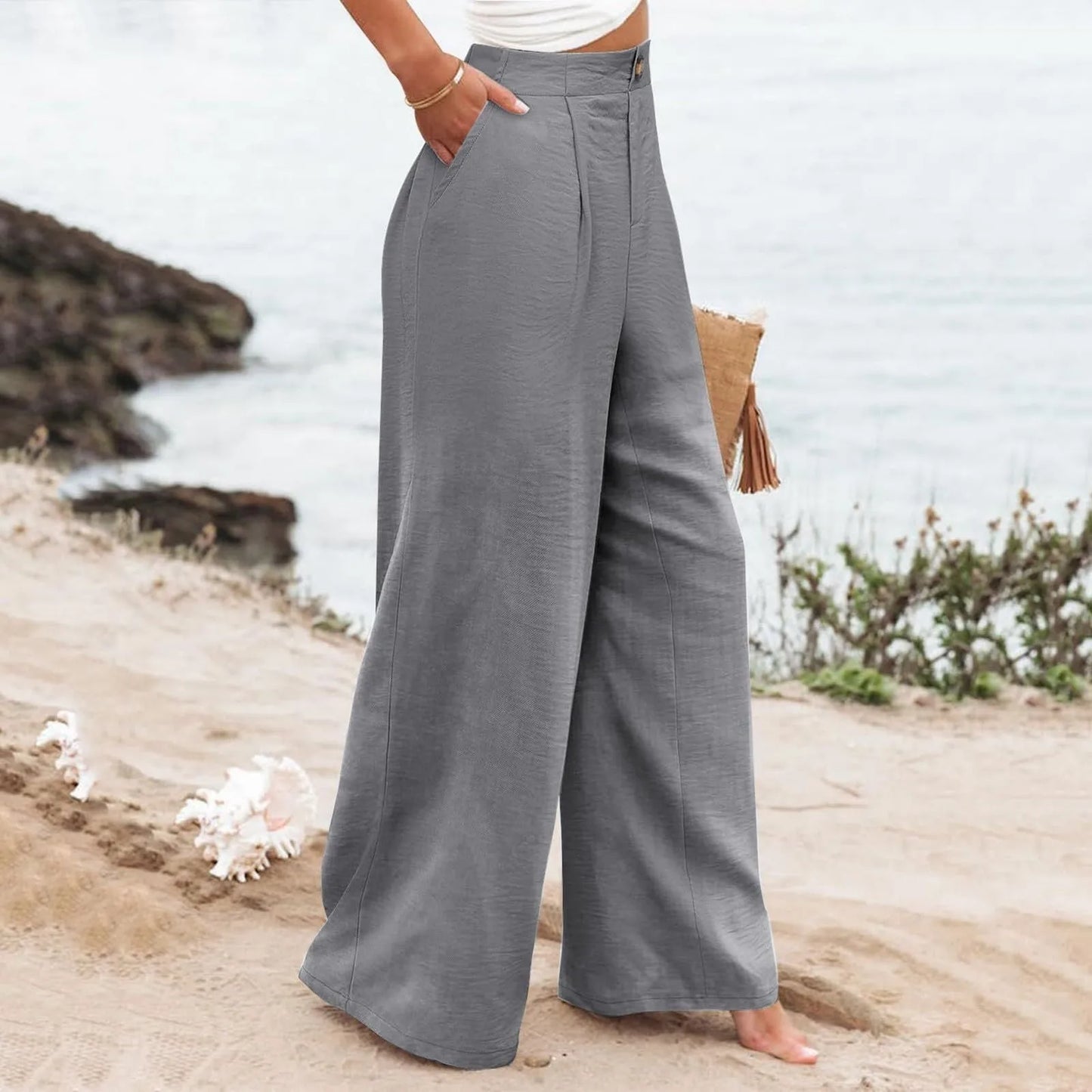 Person wearing gray wide-leg pants standing on a beach with ocean and rocks in the background.